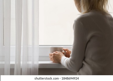 Sad adult woman sitting at window and holding mug in hands. Looking out from home. Thinking about life. Back view. 