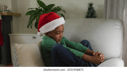 Sad african american boy wearing santa hat sitting on sofa at christmas time. christmas, festivity and tradition at home. - Powered by Shutterstock