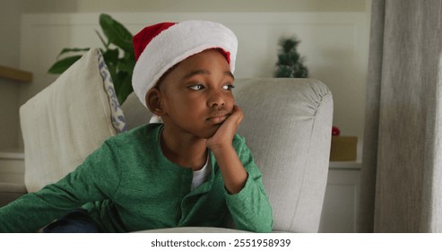 Sad african american boy wearing santa hat sitting on sofa at christmas time. christmas, festivity and tradition at home. - Powered by Shutterstock