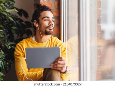 Sad African American man   listening to music from tablet  while resting near window in morning at home - Powered by Shutterstock