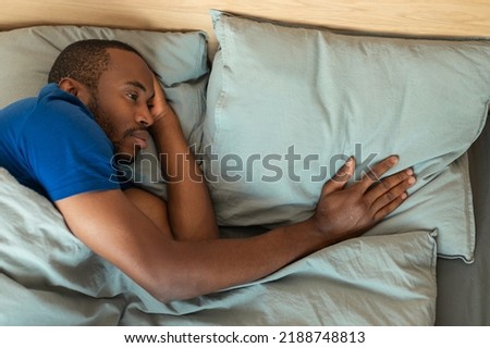 Sad African American Man Touching Pillow Suffering From Loneliness After Breakup Lying In Bed At Home. Lonely Widower Having Depression Experiencing Loss. Above View Shot.