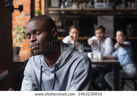 Sad african American millennial guy outcast sit alone suffering from bullying from other people in coffeehouse, depressed black man loner isolated from friends having personal psychological problems