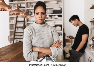 Sad African American Woman Holding Hands Together Thoughtfully Looking In Camera With Asian Man With Cellphone On Background. Young International Couple In Quarrel Spending Time On Kitchen At Home