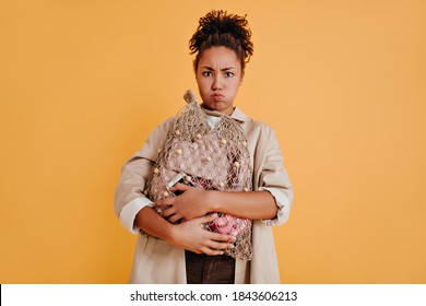 Sad African American Woman Holding String Bag. Front View Of Funny Black Girl In Trench Coat Isolated On Yellow Background.