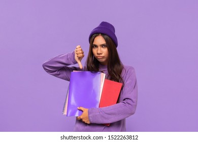 Sad African-american Teenager In A Hat And Sweater Holding Folders And Showing Thumb Down Standing Isolated Over Purple Background. Concept Of Evaluation.
