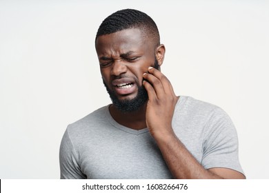 Sad afro man having gum problems, touching his cheek, white studio background, tooth disease concept