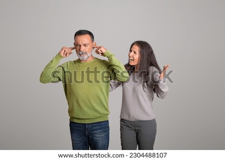 Sad angry european old wife yells at husband, man covers ears with hands on gray studio background. People emotions, stress and problems in relationships, quarrel, scandal