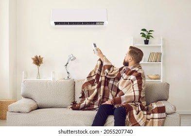 Sad Angry Man Sitting On Sofa Trying To Warm Up Under Plaid While Freezing In Cold Living Room With Modern Air Conditioner Turned On. Young Guy Having Trouble Adjusting AC System Temperature At Home