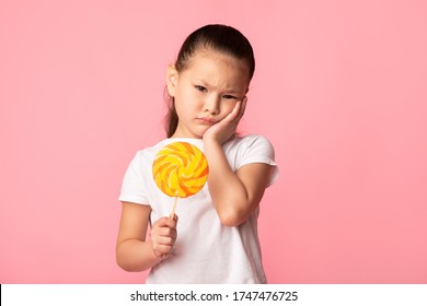 Sad Asian Child Holding Colorful Lollipop And Touching Her Cheek, Isolated Over Pink Studio Wall