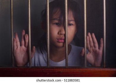 Sad Asian Child Standing Behind The Wire Screen Window In Low Key.  Unhappy Kid Alone At Home. Upset In The Dark Room