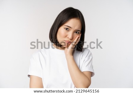 Sad asian girl looking upset and lonely, sulking and frowning, standing against white background in casual tshirt