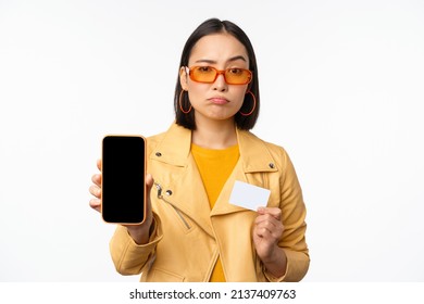Sad asian girl in sunglasses, showing smartphone app interface, credit card, looking disappointed, standing over white background