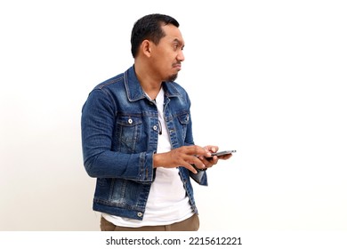 Sad Asian Man Standing While Holding A Cell Phone And Looking Beside Him On Empty Space. Isolated On White Background