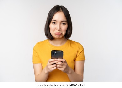 Sad Asian Woman Holding Smartphone, Looking Upset With Regret, Standing In Yellow Tshirt Against White Background