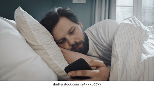 Sad attractive young man lying in bed, texting on mobile phone. Write messages with smartphone, use apps to communicate. - Powered by Shutterstock