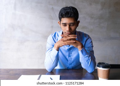 Sad Author Thinking Over Article, But Feeling Writers Block. Upset Young Indian Leaning Chin On Clasped Hands And Reflecting At Coffee Shop. Creative Crisis And Coffee Break Concept