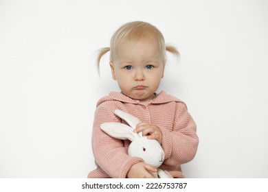 Sad baby girl play with stuffed animal isolated on white. Toddler with upset facial expression hold in hands teddy bunny. Blonde haired child in pink clothes with paschal rabbit. Easter. Holiday gift.