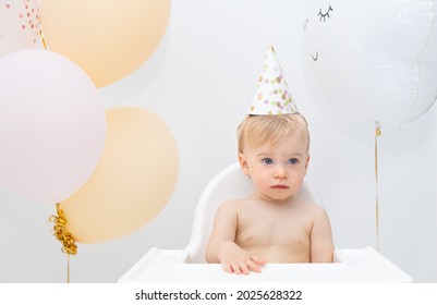 Sad Beautiful Cute Caucasian Naked Baby Sitting At Chair Among Balloons, Wearing Birthday Hat. Disappointed Kid At Celebration Party.