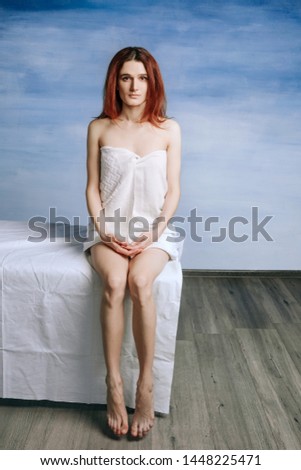 A sad beautiful young woman is sitting on a couch in a towel awaiting a doctor's appointment on a blue background.	