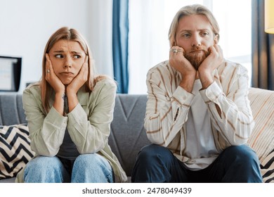 Sad, bored couple sits on a cozy sofa at home, showing signs of relationship struggles through their tired, frustrated body language