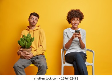 Sad Bored Man Looks At His Technology Addicted Partner Needs Lively Communication Sits On Chair Wears Sweatshirt Holds Pot Of Cactus. Happy African American Woman Uses Mobile Phone For Chatting