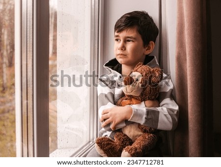 A sad boy looks out the window at the rainy autumn weather, hugging a teddy bear.