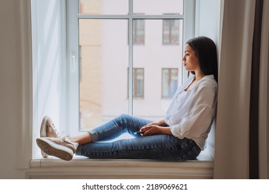 Sad Brazilian Young Woman Sitting On Windowsill In White Shirt And Blue Jeans Frustrated After Divorce, Thinking About Future Holding Phone At Home, Sunny Day. Pretty African American Girl In Troubles