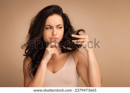 Sad brunette young woman holding her long hair split ends in hand and touching chin isolated on beige background, need for mask treatment, diet correction, balm, vitamins, copy space