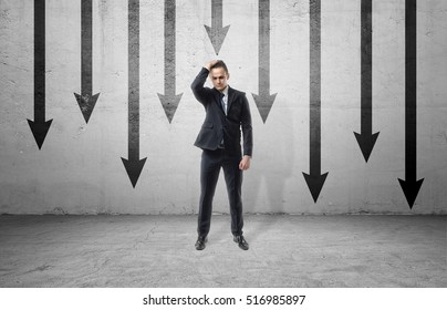 A sad businessman in a frustrated pose, standing on the background of many black arrows pointing down, painted on a grey wall. Business and management. Failures and problems. Poses and gesrures.