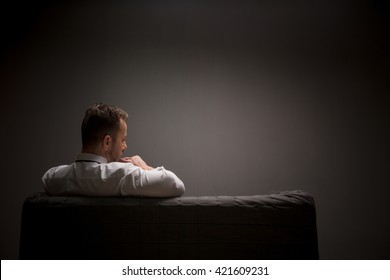 Sad Businessman Sitting On Comfortable Sofa After Hard-working Day And Continues Thinking About Work. Studio Shot. Back View.