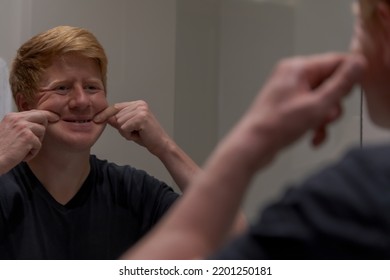 Sad caucasian redhead man in black t-shirt forces himself to smile by pulling his cheeks to the sides with his hands in front of mirror. Selective focus. Mental health theme.