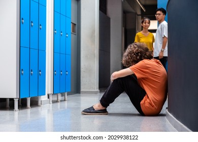 Sad caucasian teen boy sitting on floor in high school corridor while his classmates observe him. Copy space. Bullying consequences concept. Juvenile Depression - Shutterstock ID 2029938122