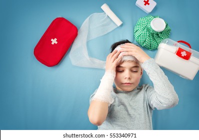 Sad Child In Bandages Lying In Bed. Wounded Boy On Blue Background With First Aid Accessories.