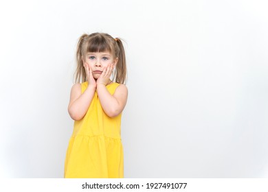 Sad Child Girl In A Yellow Dress Posing On A Gray Background, Hands Near The Face