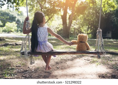 Sad Child Holding Hand With Teddy Bear On Wooden Swing In Park. Asian Kid Sitting With Best Friends Forever, Sad Moment. Teddybear Is A Gift, Toy And Best Friend For Children. Friendship Concept