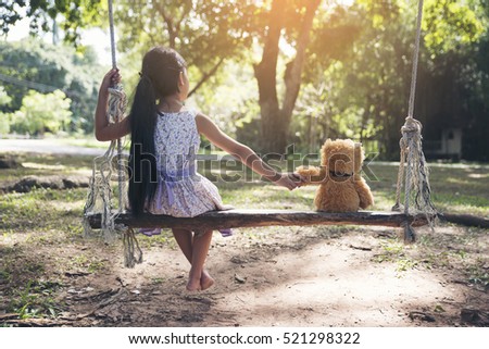 Sad child holding hand with teddy bear on wooden swing in park. Asian kid sitting with best friends forever, sad moment. Teddybear is a gift, toy and best friend for children. Friendship concept