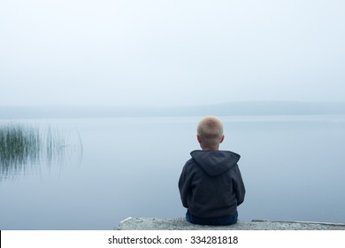 Sad Child Sitting Alone By Lake In A Foggy Day, Back View