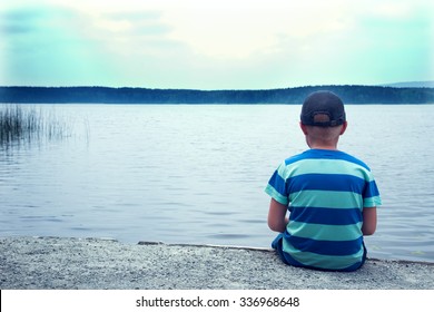 sad child sitting alone at the lake on a cloudy day, back view