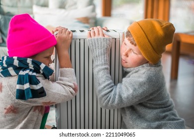 Sad Children Hugs Radiator. Kid In Hat And Warm Sweater Near Home Heater. Winter, Heating. Sad Boy And Girl. Energy Crisis In The Winter Cold Season. Restrictions And Savings Of Gas And Electricity