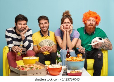 Sad Company Of Friends Feel Bored As Watch Not Interesting Match On Television, Surrounded With Different Delicious Snacks, Happy Guy Holds Money, Makes Bet On Game, Pose At Comfortbale Sofa