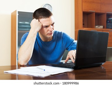 Sad Confused Man Looking At Laptop Sitting By Table In Home Interior