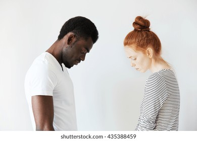 Sad couple looking down with their heads bowed in front of each other. Side view portrait of two sorrowful people: young Caucasian redhead girl and Afro-American melancholic man. Negative emotions.
