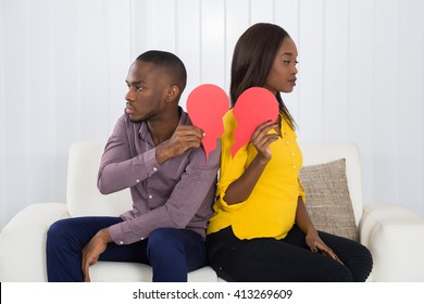 Sad Couple Sitting Back To Back On Sofa Holding Red Broken Heart