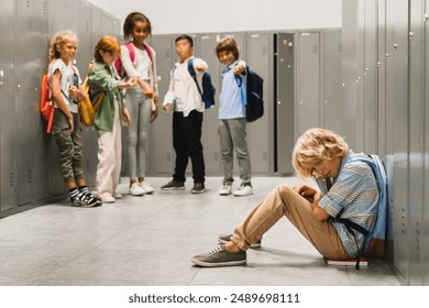 Sad crying schoolboy pupil sitting on the floor at the school hall while his classmates teenagers laughing at him, bullying and torturing him. Social exclusion problem - Powered by Shutterstock