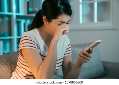 sad crying teen girl checking phone sitting on sofa in dark living room at home. asian woman overworked nearsighted looking for long time on smartphone screen rubbing painful eyes in late night