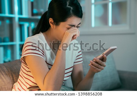 sad crying teen girl checking phone sitting on sofa in dark living room at home. asian woman overworked nearsighted looking for long time on smartphone screen rubbing painful eyes in late night