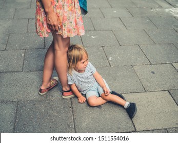 A sad crying toddler is sitting in the street by his mother's feet