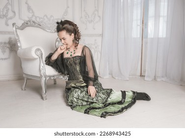 Sad crying woman wearing green medieval vintage Victorian Style dress sitting on the floor near armchair - Shutterstock ID 2268943425