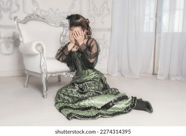 Sad crying woman wearing green medieval vintage Victorian Style dress sitting on the floor near armchair - Shutterstock ID 2274314395