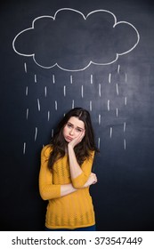 Sad Cute Young Woman Standing Under Raincloud And Rain Drawn Above Her On A Blackboard Background